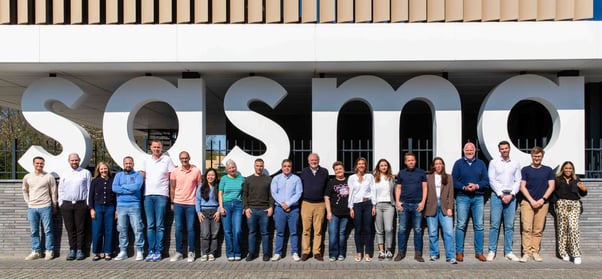 A line of people stand outside of a building in front of a large logo reading 'Sasma'