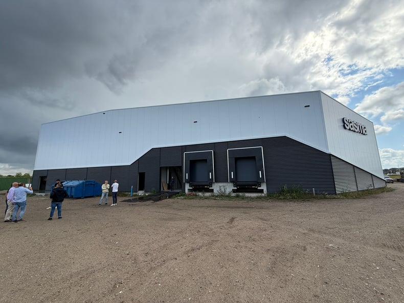 A large black and white warehouse building against a cloudy sky. The building has the logo Sasma on it