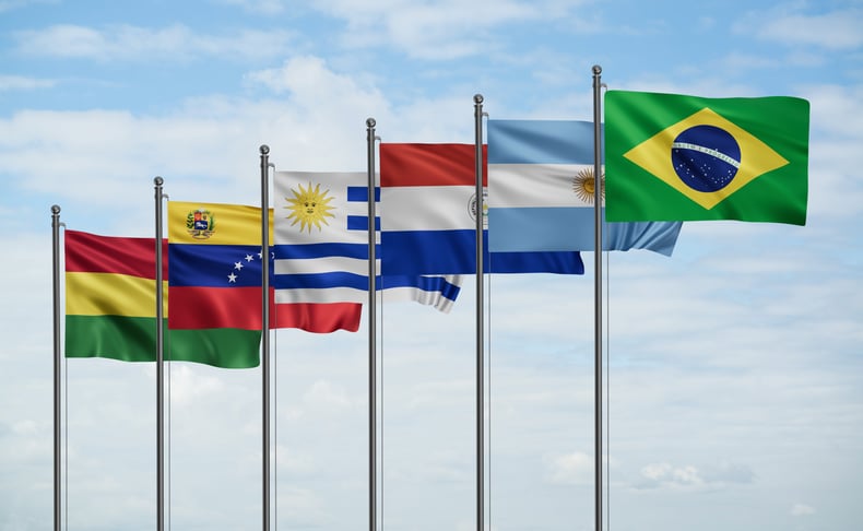 Six South American flags fly from flagpoles against a partly cloudy sky